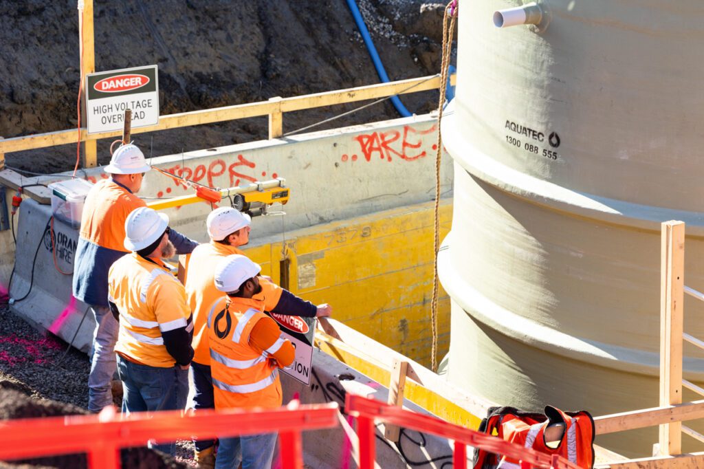 Work men wearing high vis clothing and a hard hats looking at a large tank being lowered into the ground