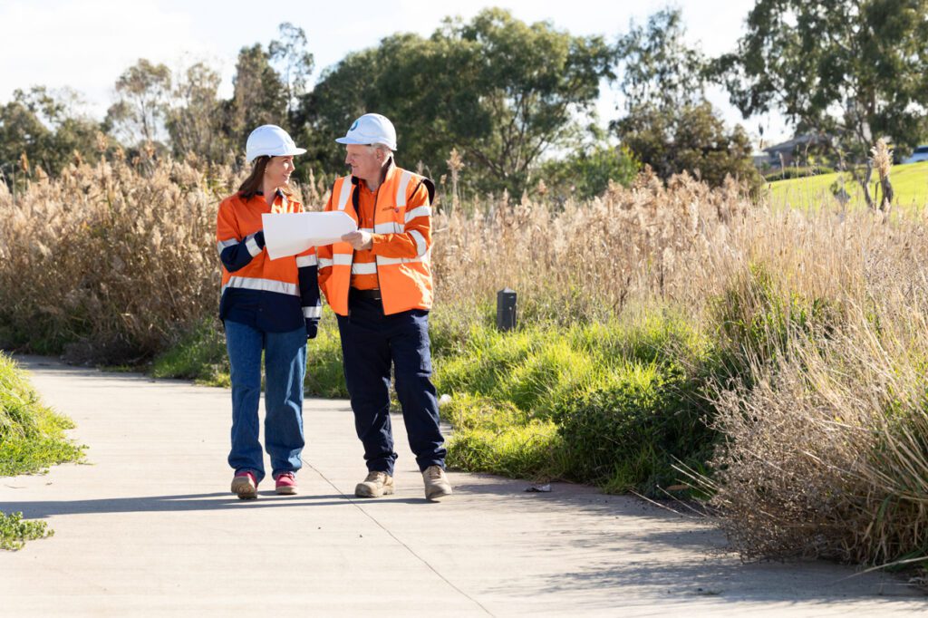 A male and a female wearing high vis clothing and hard hats looking over work plans as they walk along a path