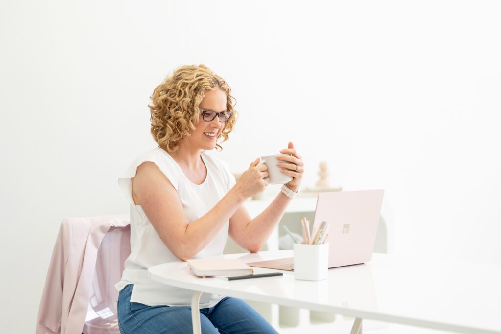 Photo of a female entrepreneur sitting at her desk, looking at her laptop while holding a cup of coffee and she is wearing a white top and has her eye glasses on