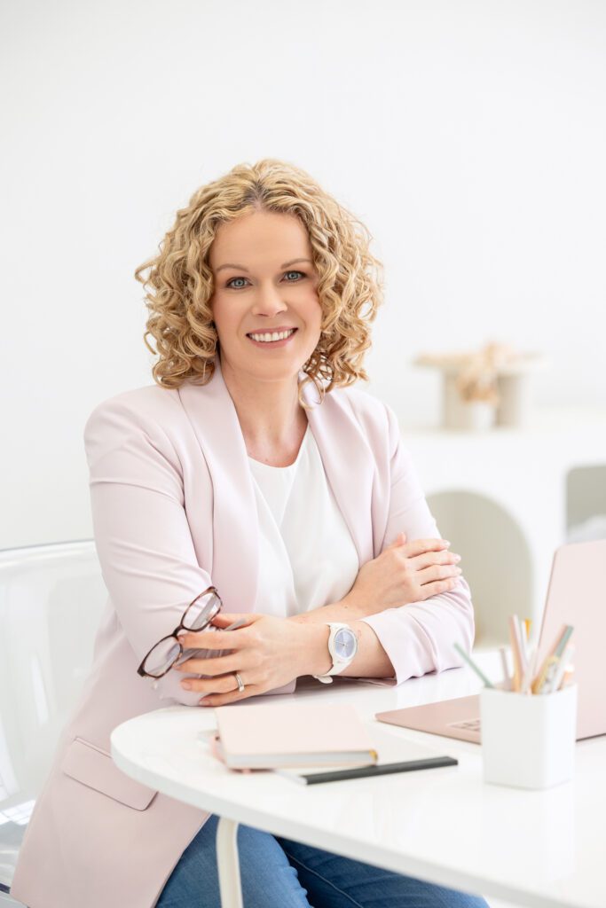 Portrait of a female entrepreneur sitting at her desk, with her laptop, notebooks, coffee cup and pen holder in the foreground, she is posed with her arms cross in front of her leaning on the desk and holding her eye glasses in her left hand, she is wearing a white top and pale pink blazer