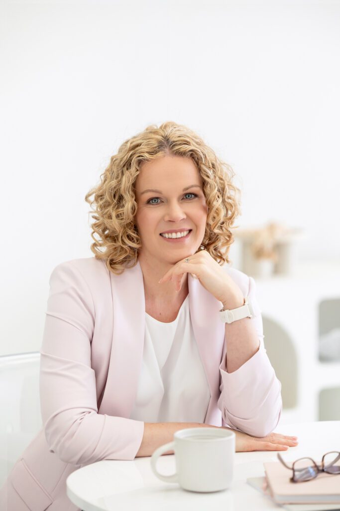 Portrait of a female entrepreneur sitting at her desk and leaning her chin on one of her hands while wearing a white top and pale pink blazer