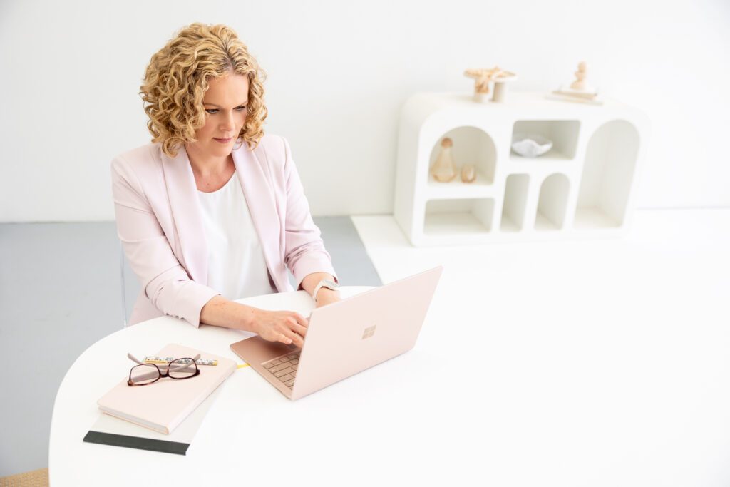 Photo of a female entrepreneur taken from an elevated angle while she sits at her desk and types on her laptop, she is wearing a white top and pale pink blazer