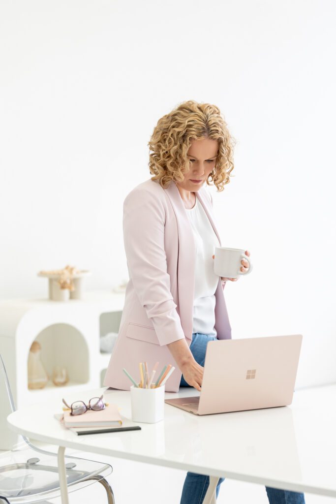 Photo of a female entrepreneur leaning against her desk and looking down and checking her laptop while holding a cup of coffee, she is wearing a white top, blue jeans and a pink blazer