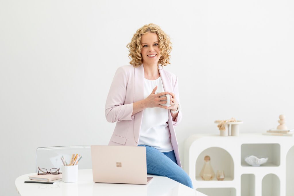 Portrait of a female entrepreneur leaning on her desk which has her laptop on it while holding a cup of coffee, she is wearing a white top, blue jeans and a pink blazer