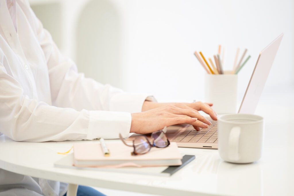 Close up photo of hands typing on a laptop keyboard with a notebook, pen, eye glasses and cup in the foreground.