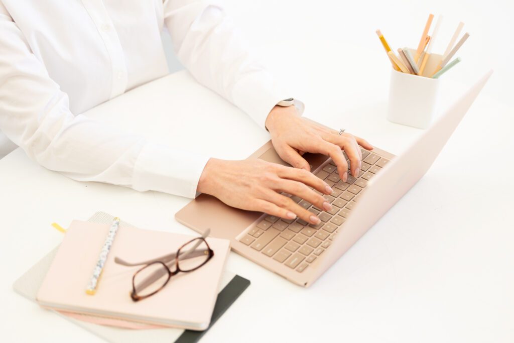 Photo of a desk with hands typing on a laptop keyboard with a notebook and eye glasses in the foreground