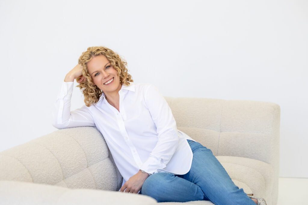 Photo of a female entrepreneur looking relaxed and leaning into the side of a couch couch while wearing a white linen shirt and blue jeans