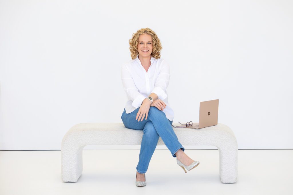Full length portrait of a female entrepreneur shot in a studio while sitting on a bench seat with her open laptop beside her and wearing a white linen shirt and blue jeans
