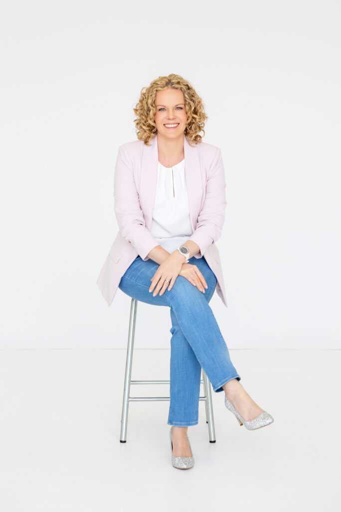 Full length portrait of a female entrepreneur shot in a studio while sitting on a stool and wearing a white shirt, blue jeans and a pale pink blazer