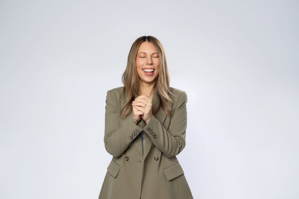 Portrait of a female business owner wearing a khaki green pant suit standing in front of a blank backdrop with her eyes closed laughing