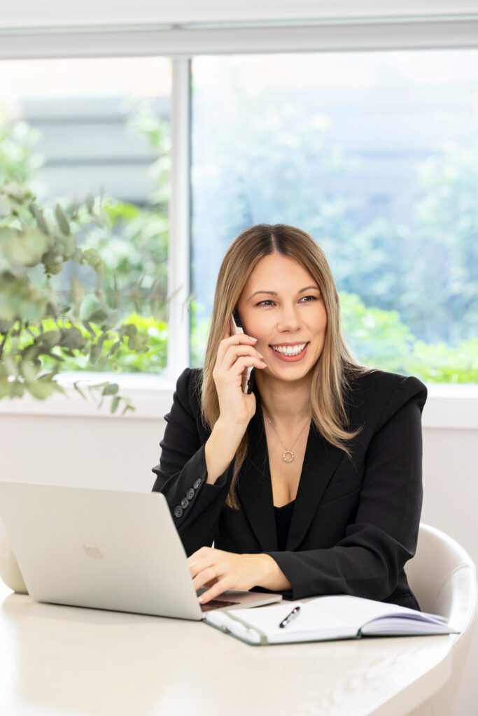 A female business owner wearing a black blazer sitting at a table with her laptop open and talking into her mobile phone