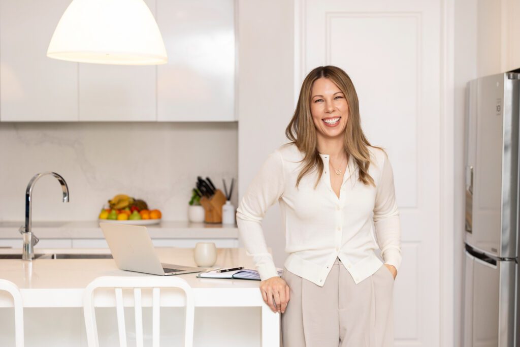 A female business owner wearing a neutral toned cardigan and pants standing at the end of her kitchen bench with her laptop and notebook sitting on the bench