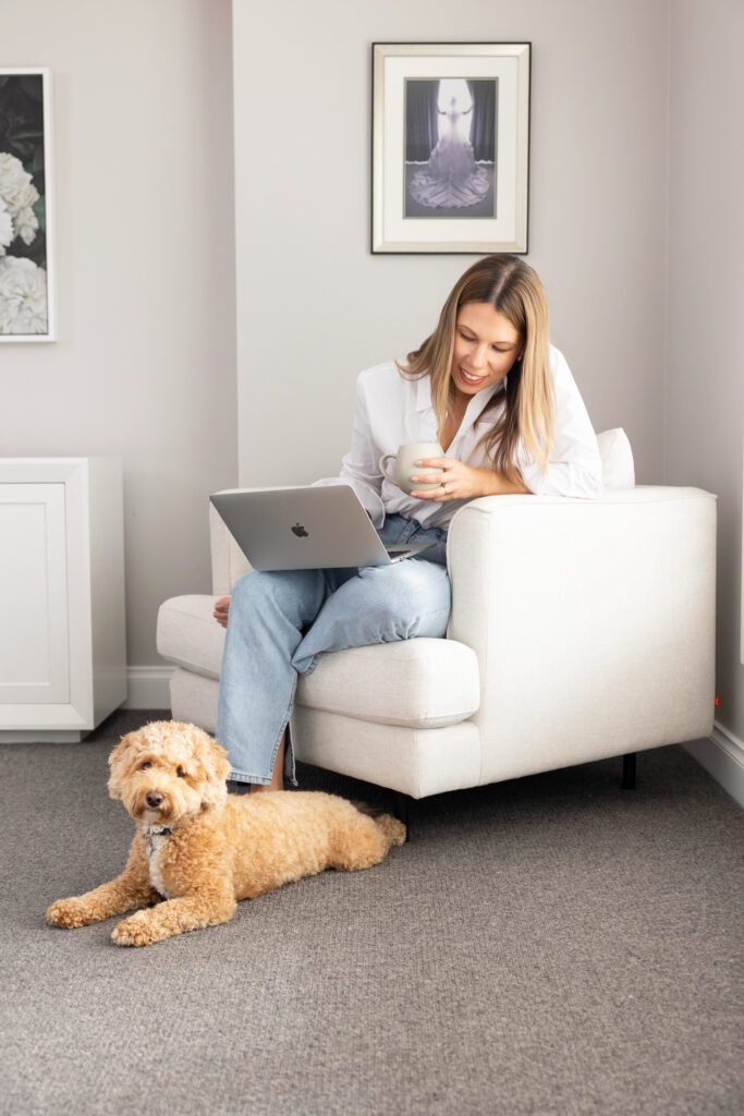 A female business owner wearing a white shirt and blue jeans sitting in a white arm chair balancing a laptop on her legs and holding a coffee cup while looking down at her dog that is laying down on the flow at her feet