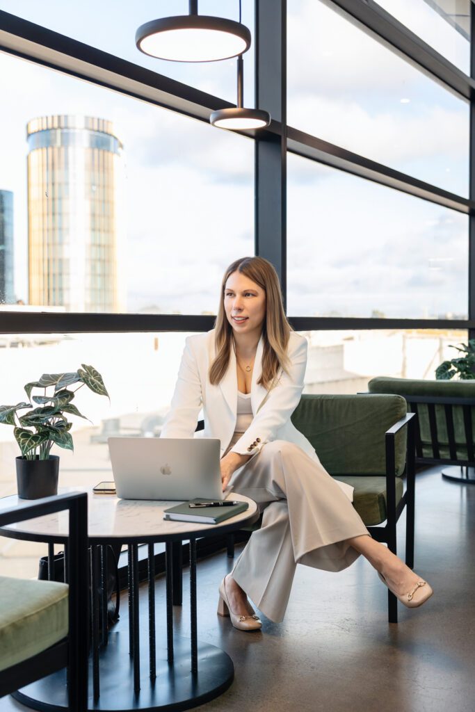 A female business owner wearing a white blazer and neutral coloured pants sitting and a table working on her laptop