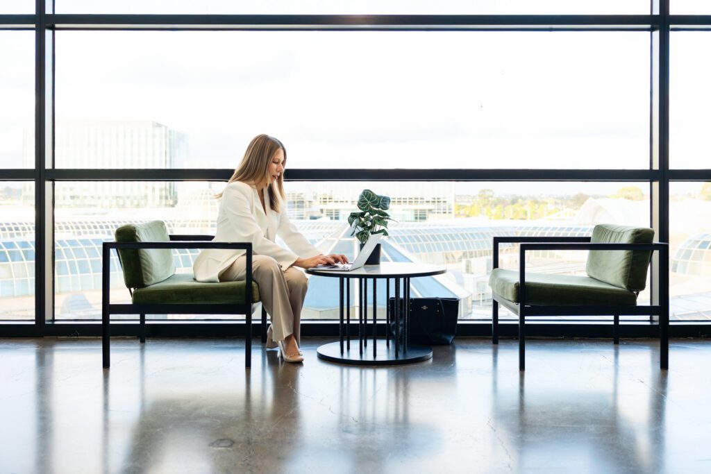 A female business owner wearing a white blazer and neutral coloured pants sitting and a table working on her laptop