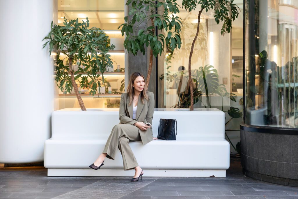A female business owner wearing khaki pant suit sitting on a bench holding her mobile phone