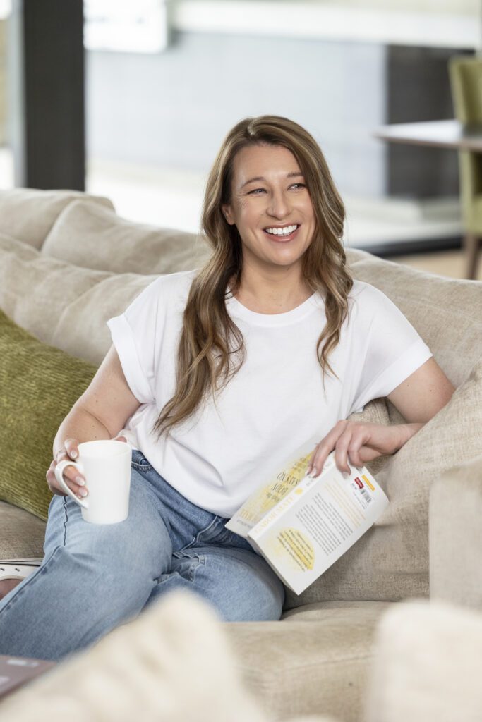 Personal Branding Photography of a female psychologist wearing a white t-shirt and jeans sitting on her couch holding a book and a cup of coffee